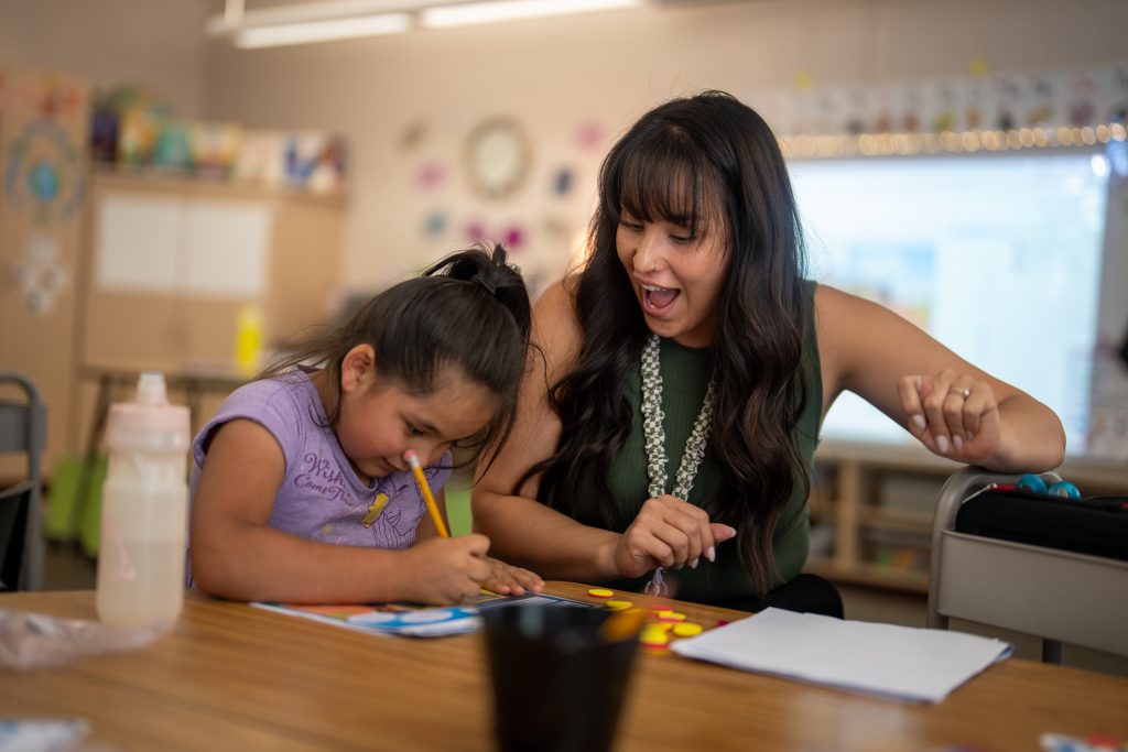 A teacher and a young girl engage in a creative activity at a table, both smiling and focused on their work.