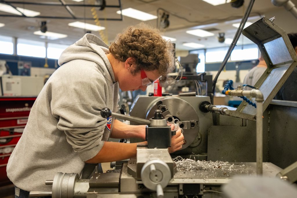 A student works on a lathe machine in a workshop, focusing intently while wearing safety glasses. Shavings scatter around.